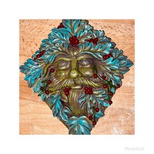 May include: A green man's face with a beard, surrounded by blue and green leaves and red berries. The face is made of a textured material and has a rustic look.