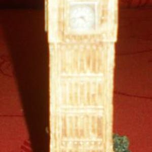 May include: A detailed miniature model of the Big Ben clock tower. The beige tower has a green roof and a clock face. The base includes green shrubbery and a blue water feature. A decorative souvenir.