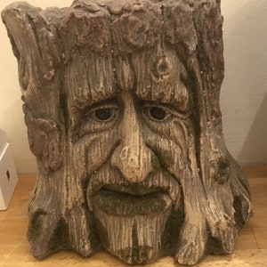 May include: A weathered, tree-stump-shaped planter with a carved face. The face has a long nose, a full mustache and beard, and deep-set eyes. The planter is primarily brown with hints of green moss.
