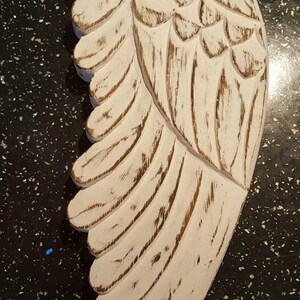 May include: White wooden angel wing with a distressed finish. The wing has a carved design and is attached to a twine string.