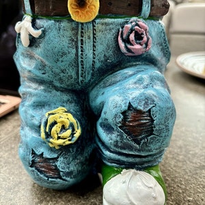 Op de afbeelding: Een blauwe denim plantenbak in de vorm van een spijkerbroek met een bruine riem, een gele bloem, een roze bloem en een gele bloem aan de voorkant. De spijkerbroek heeft een gescheurde knie en een witte schoen.