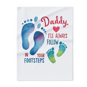 Może przedstawiać: Biała tkanina z akwarelowym wzorem przedstawiającym niebieskie i zielone odciski stóp. Tekst brzmi "Daddy, I'll always follow in your footsteps" w kolorze czerwonym i niebieskim, z czerwonym symbolem serca.