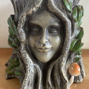 May include: A ceramic planter shaped like a tree trunk with a woman's face carved into it. The planter is painted in shades of brown and green, with a small orange mushroom at the base.