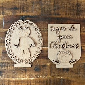 May include: Two wooden Christmas decorations. One is an oval-shaped gingerbread man with a decorative border. The other is a rectangular sign with the words "Sugar & Spice Christmas" and a gingerbread man.