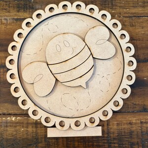 May include: A wooden decorative piece featuring a cartoon bee design. The bee has striped detailing and wings, set within a scalloped circle. The piece is made of unfinished wood and includes a stand for display.