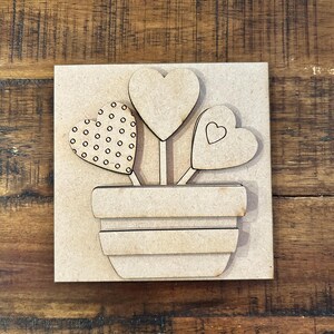 May include: A wooden craft featuring a flower pot with three heart-shaped flowers. One heart has a dotted pattern, another has a heart outline, and the third is a solid heart. The craft is made of unfinished wood.