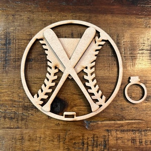 May include: A baseball-themed wooden cutout featuring crossed bats, a baseball, and a ring. The baseball design includes stitching and a circular frame. The ring is separate. The cutout is made of light brown wood.