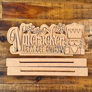 May include: Wooden Christmas nutcracker sign with the text "Nutcracker Let's Get Crackin'" and a nutcracker figure with a crown and beard. The sign is on a wooden background.
