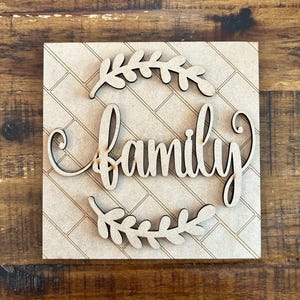 Może przedstawiać: Drewniany znak z napisem "rodzina" w kursywie, otoczony wzorem wieńca laurowego. Znak umieszczony jest na jasnobrązowym, teksturowanym tle z wzorem ceglanym. Dekoracyjny przedmiot.