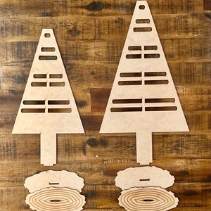 May include: Two unfinished wooden Christmas tree ornaments with a layered, circular base. The trees have a simple design with a pointed top and horizontal lines.