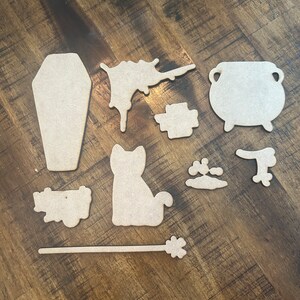May include: A set of nine wooden Halloween-themed cutouts, including a coffin, a cauldron, a cat, a wand, a splattered paint design, a paw print, a broom, and two other shapes.