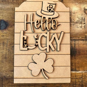 May include: Wooden sign with the words "Hello Lucky" in a whimsical font. The sign features a shamrock, a horseshoe, and a leprechaun hat.