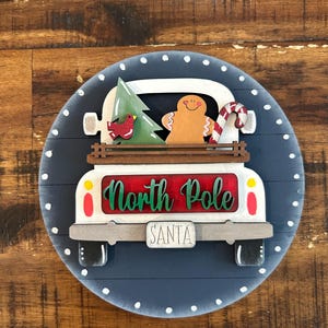 Może przedstawiać: Okrągła, drewniana dekoracja świąteczna z białym samochodem z czerwonym napisem "North Pole". Na pace znajduje się zielone drzewko, piernikowy ludzik i laska cukrowa. Tło to ciemnoniebieskie koło z białymi kropkami.