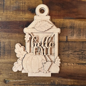 Puede incluir: Decoración de madera en forma de linterna con las palabras "hello FALL" recortadas. La linterna presenta una calabaza, hojas y una pequeña baya. La decoración tiene un agujero en la parte superior para colgar y es de madera sin terminar.