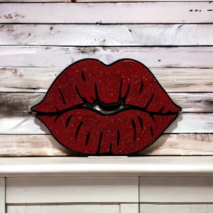 May include: Red glitter lips with a black outline, displayed on a white wood background.