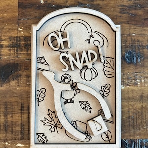 May include: A wooden sign with the words "OH SNAP!" and Thanksgiving-themed illustrations. The sign features a turkey, wishbone, pumpkin, and fall leaves. The sign is made of light-colored wood and has a decorative border.