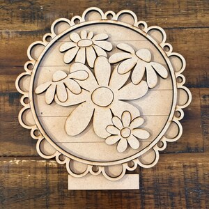 May include: A wooden floral decoration featuring a circular design with a scalloped edge. The center showcases several daisy-like flowers. The piece is supported by a rectangular base, perfect for display on a shelf or table.