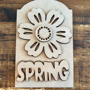 May include: Wooden craft blank with a cutout flower and the word "SPRING" in large letters. The blank is unfinished and ready to be painted or stained.