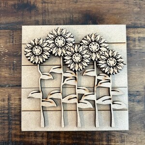 May include: A wooden sunflower art piece featuring five sunflowers with detailed petals and textured centers. The sunflowers are attached to stems with leaves, all mounted on a square, light-colored wooden base. The piece is designed with a natural, rustic aesthetic.