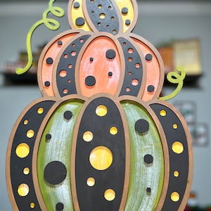 Puede incluir: Una decoración de calabaza de madera de tres niveles con un patrón de lunares verdes, negros y amarillos. La calabaza superior es amarilla con lunares negros, la calabaza del medio es naranja con lunares negros y la calabaza inferior es verde con lunares negros y amarillos.