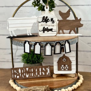 May include: A two-tiered farmhouse-style tiered tray with a wooden base and a galvanized metal top. The tray is decorated with various farmhouse-themed wooden cutouts, including a cow, a pig, a chicken, a barn, and a sign that reads "Life is better on the farm". The tray also features a wooden sign that reads "Farmhouse" and a string of milk bottle cutouts.