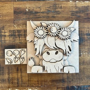 May include: Wooden Highland cow with sunflower crown and separate small wooden pieces. The cow is light brown with a detailed design. The sunflowers are intricately cut. The small pieces appear to be berries and leaves.
