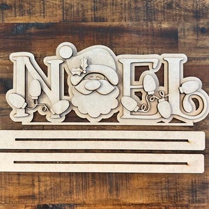 May include: Wooden craft kit featuring the word "NOEL" with a Santa Claus face in the center. The letters are decorated with string lights. The kit includes two wooden slats for hanging.