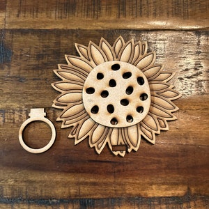 May include: A wooden sunflower ornament with a ring-shaped attachment. The sunflower has detailed petal and seed designs, with several small holes in the center. The ornament is made of light-colored wood and is likely for crafting or decoration.