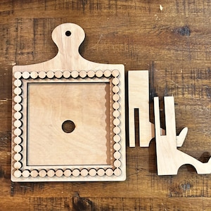 May include: A light-colored wooden square frame with a handle and decorative border, along with two support pieces. The frame has a central hole and a scalloped edge. The support pieces are designed to hold the frame upright.