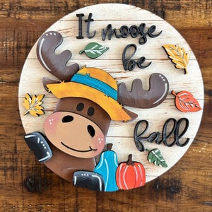 Könnte beinhalten: Ein rundes Holzschild mit einem Elch mit Hut, dem Text "It moose be Fall" und herbstlichen Dekorationen. Der Elch ist braun mit einem gelb-blauen Hut. Das Schild hat einen weißen Hintergrund mit Holzlatten.