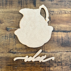 May include: A wooden cutout of a pitcher and a separate wooden cutout of the word "relax" in a cursive font. The cutouts are light brown and sit on a dark brown wooden surface.
