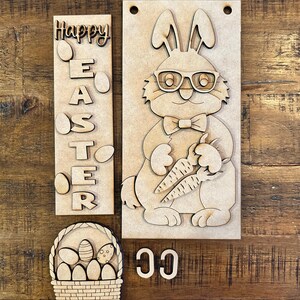 Puede incluir: Kit de manualidades de madera para hacer un conejito de Pascua, una cesta y un letrero. El conejito lleva gafas y sostiene una zanahoria. El letrero dice "Feliz Pascua" con huevos alrededor de las letras. La cesta tiene huevos dentro.
