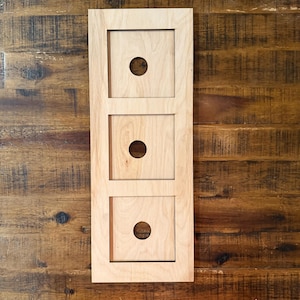 May include: A rectangular wooden wall hanging with three square cutouts, each featuring a centered circular hole. The wood has a light, natural finish, and the piece is mounted on a dark brown wooden surface.