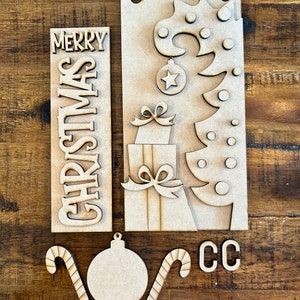 May include: Wooden Christmas craft kit with a "Merry Christmas" sign, a Christmas tree, a present, a candy cane, and an ornament. The kit also includes the letters "CC".