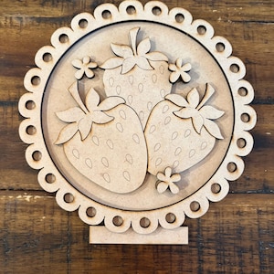 May include: A wooden strawberry-themed decorative piece. Three strawberries are centered within a scalloped circle, with small flower accents. The piece is made of light brown wood and stands on a rectangular base.