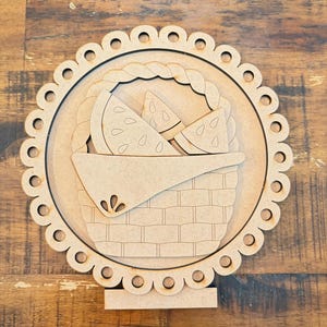 May include: A wooden watermelon picnic basket cutout. The basket is filled with watermelon slices and is surrounded by a scalloped edge. The cutout is made of light brown wood and is suitable for crafts or home decor.