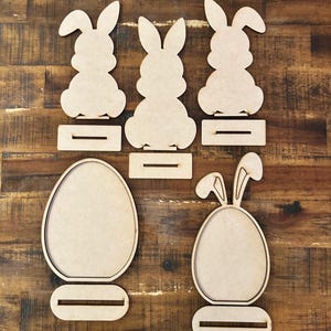 May include: Wooden Easter decorations featuring bunny and egg shapes. The set includes three bunny figures and two egg-shaped frames, all with a slot for display. The unfinished wood allows for customization.