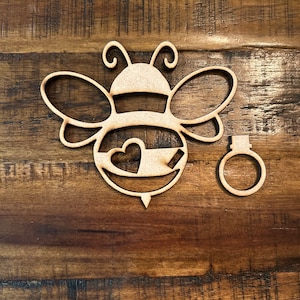 May include: Wooden cutout of a bee with a heart in its body and a separate wooden ring. The bee is brown and the heart is a lighter brown.
