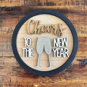 Può includere: Un cartello rotondo in legno con la scritta "Cheers to the New Year". Il design include calici di champagne e un bordo nero. Una decorazione festosa per i festeggiamenti di Capodanno.