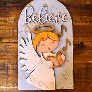 May include: Wooden sign featuring a cartoon angel with orange hair, a halo, and wings, singing from a book. The word "Believe" is written above the angel. The background is light blue with musical notes.