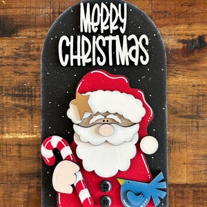 Può includere: Decorazione natalizia in legno raffigurante Babbo Natale con barba bianca, abito rosso e bastoncino di zucchero. Le parole "Merry Christmas" sono in scrittura bianca su sfondo nero con macchie bianche. È presente anche un uccellino blu.