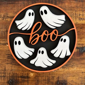 Może przedstawiać: Okrągła dekoracja na Halloween z pięcioma białymi postaciami duchów na czarnym tle. Słowo "boo" jest napisane pomarańczową czcionką. Dekoracja ma pomarańczową obwódkę.