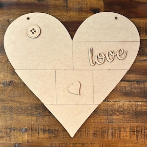 May include: A heart-shaped wooden craft with segmented sections. Features a button, the word "love", and a small heart cutout. The craft is made of light brown wood and has a hole at the top for hanging.