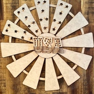 May include: A wooden craft project in the shape of a windmill with the word "USA" in the center. The windmill has a circle of stars around the center and wooden blades.