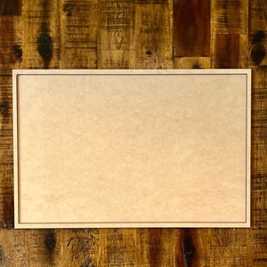 May include: A blank, light brown, rectangular picture frame with a thin black border. The frame is sitting on a wooden surface.