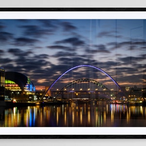 May include: A night-time view of a city skyline with a large arch bridge illuminated in purple and white lights. The bridge is reflected in the water below, along with the lights of the city. The sky is a dark blue with clouds.