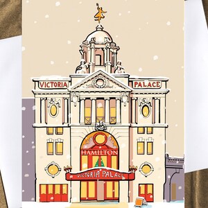 May include: A greeting card featuring an illustration of the Victoria Palace Theatre in London during a snowfall. The card has the words "VICTORIA PALACE" and "HAMILTON" on the front. A small sign is in front of the theater.