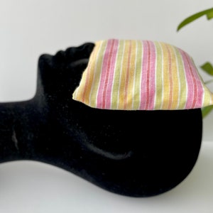 May include: A striped eye pillow with pink, yellow, and green stripes. The pillow is made of a soft, light fabric and is perfect for relaxation.