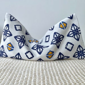 May include: A white decorative pillow with a geometric pattern of navy blue and yellow shapes. The pillow is folded in a triangular shape, showcasing the fabric's design. The pillow is on a textured, off-white surface.