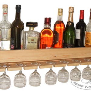 May include: A wooden wall-mounted wine rack with a shelf for bottles and a lower shelf for hanging wine glasses. The rack is made of natural wood and has a light brown finish. It is holding various bottles of wine and liquor, including a bottle of Disaronno.